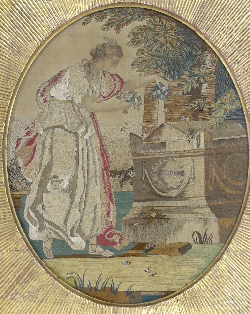 A gilt framed Regency oval silkwork picture of a classical maiden scattering flowers at a tomb, labelled Cowper, probably commemorating the death of the poet William Cowper in 1800, 30x25cm